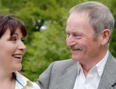 Lorraine Kelly/Instagram A smiling woman and an older man with a moustache look at each other outdoors, both wearing smart jackets, with green foliage blurred in the background.