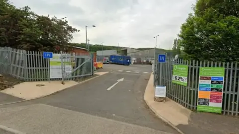 Google The entrance to a recycling facility with metal fencing, “IN” signs on both sides of the gate, and several information boards. A green sign promotes local recycling, while modern green‑and‑grey buildings stand behind the fenced area under a clear blue sky.
