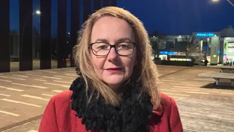 Sam Read/BBC Lou Cory is standing on a square in Corby in the early evening. She has fair hair and is wearing glasses, a red coat and a black scarf. Shops are lit up in the background