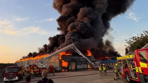 Essex County Fire and Rescue Service Black smoke billows from a warehouse fire in Essex. There are fire engines and fire fighters in the foreground. 