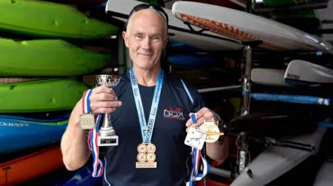 St Monica Trust A man called Jules Taylor stands inside a shed lined with canoes and kayaks, holding various medals he won for becoming Quadrathlon World Champion for his age group. He is smiling and is wearing a dark blue T-shirt and has a pair of sunglasses perched on his head