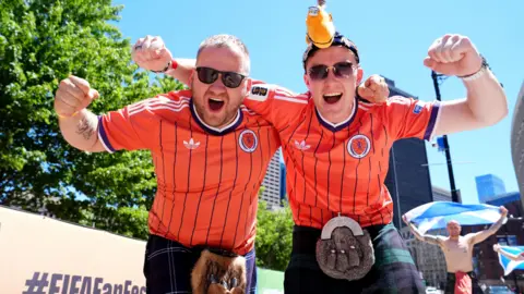 Two fans outside the stadium in Boston. They are wearing Scotland away strips and kilts, as well as sunglasses, and have their arms raised.