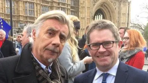 Sex Pistol Glen Matlock and former Cardiff West MP Kevin Brennan at a demonstration surrounded by other people outside the Houses of Parliament. Both are dressed smartly and looking at the camera