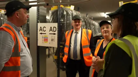 The Lib Dem leader Sir Ed Davey, standing in front of a row of clothes in plastic packaging on what appears to be a factory floor. He is wearing a cap, a suit and tie, and an orange high-vis jacket. He is listening while three other people talk.