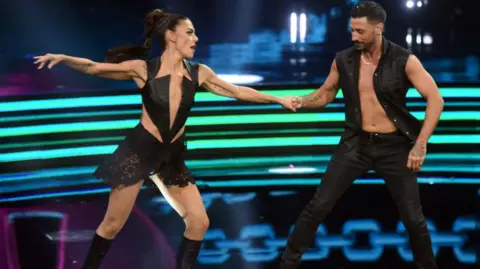 Getty Images Bianca Guaccero and Giovanni Pernice dressed in black outfits hold hands mid-dance routine with green and blue neon lights in the background.