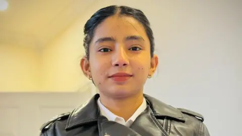 A young woman with dark brown hair tied back is wearing a white shirt and black leather jacket and standing indoors. She is looking at the camera and has a hoop earring in each ear.
