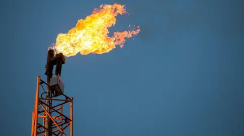An orange tower on the left of the picture, a orange flame is being emitted with black smoke trailing left to right. The tower stands against a blue sky. The flame is produced by flaring from oil production