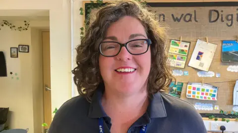 A woman with a curly dark brown and grey bob stands in a classroom with a wall display behind her. She is wearing a black polo shirt and black framed glasses and is wearing red lipstick. She has a small silver hoop nose-ring and is wearing a lanyard with a ribbon that reads 'staff'