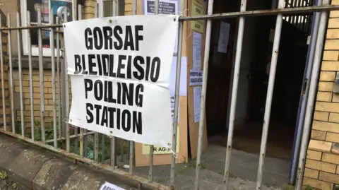 BBC Bilingual sign saying "polling station/gorsaf pleidleisio" attached to railing outside a village hall.