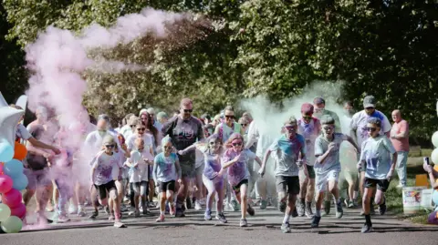 A group of men, women and children running together towards the camera. They are wearing sports clothing and googles, while pink and blue powder surrounds them.