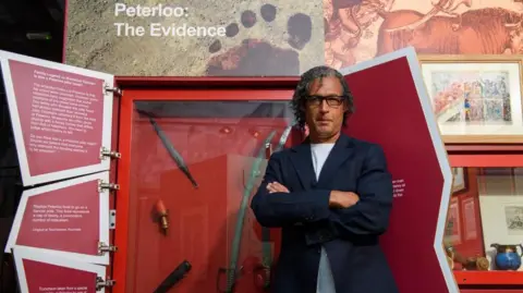 PA Media/Jon Parker Lee/Findmypast A man with short grey and black hair and wearing black framed glasses, a dark coloured jacket and white T-shirt has his arms folded while standing next to a Peterloo Massacre display. The display of weapons and other artefacts has a sign saying 'Peterloo: The evidence' above it. 