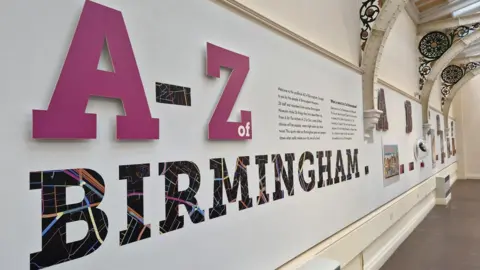 Ruth Millington Close-up view of a museum wall display reading “A–Z of Birmingham,” with large pink letters and patterned typography, alongside explanatory text panels and exhibits under decorative arched ironwork and a glass ceiling.
