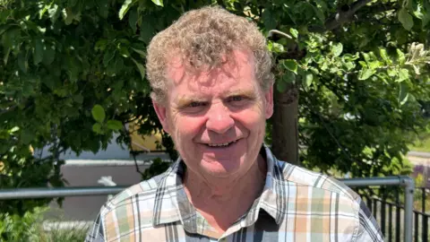 BBC / Gavin Kermack A man with short light brown curly hair, wearing a check-patterned shirt, standing in front of a leafy green tree on a sunny day.