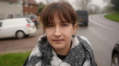 Qays Najm/BBC Jessica Smith standing outside by the side of the road. She is looking directly at the camera. She has brunette hair with a full fringe and is wearing a white and grey scarf round her neck.