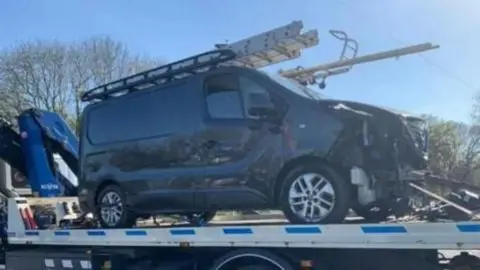 A grey Nissan van, with severe damage to its front,  loaded on to a tow truck 