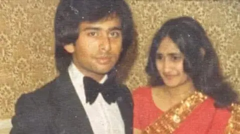 George Miah The image appears to be an older, slightly faded photograph showing two people standing indoors. One man is wearing a black formal suit with a white shirt and a bow tie, while the other is dressed in a bright red, patterned outfit with a decorative border. The setting looks like a domestic or reception room, with patterned wallpaper in the background and a chair partially visible to the side. The photograph has a vintage look, with noticeable grain and wear consistent with an image taken several decades ago.