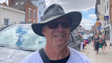 A photo of a white man wearing sun glasses and a wide brimmed hat and a white t-shirt. He is stood in front of a car with a street to his right, with two women walking towards him