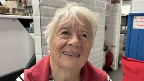 An elderly woman with short white hair smiles with her top row of teeth showing. She is stood in front of a white tiled wall. She is wearing a red gilet.