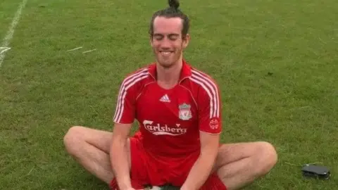 Family flyer Liam McGenity sat cross-legged on a football pitch wearing his Liverpool football team kit