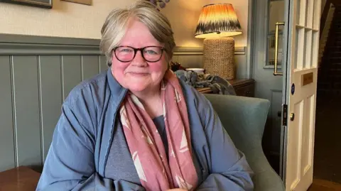 Vikki Irwin/BBC A woman sitting in a cosy room with wood paneled walls which are painted grey-green. She is wearing a light blue jacket and top with a pink scarf with white sailing boats as a pattern.