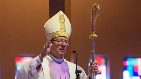 John Sherrington wears an ornately decorated white and gold mitre and a white and lilac robe. He holds a long, thin staff in his left hand known as a crosier.