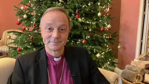 The Bishop of Exeter, Dr Mike Harrison, sat on a cream sofa. Behind him is a Christmas tree with red and gold baubles and strong lights. To the right of him is chopped up wood. He is wearing a purple shirt and black blazer. 