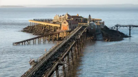 Birnbeck Pier in Weston-super-Mare. The pier is damaged and in disrepair, with a heavily damaged wooden walkway and dilapidated buildings.