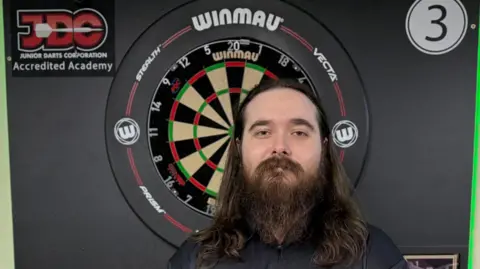 Dan McNamara, who has long hair and a beard, stands in front of a dartboard