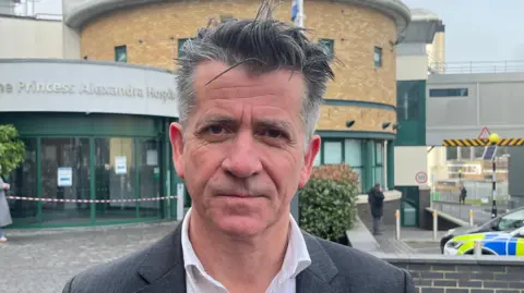 Michael Meredith, who is wearing a white shirt and grey suit jacket. He has short grey hair and is stood in front of the hospital building with a serious look on his face.