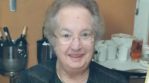 Pamela Kinrade has short wavy grey hair and wears glasses. She is smiling, sat in front of a table with bottles of wine to the left and white creamic mugs to the right.
