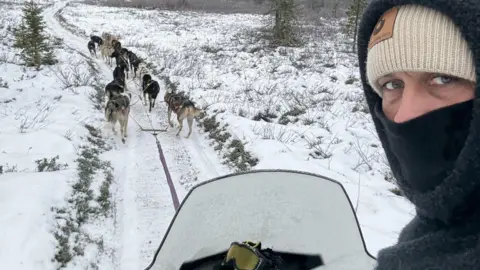Danny Welch turning to look over his shoulder, with a grey fleece hood up covering a cream wool hat and a black balaclava pulled up to his nose obscuring most of his face. He is sitting on a sledge with a glass screen in a snowy landscape and ahead of him are a team of huskies pulling the sledge along a snowy track. 