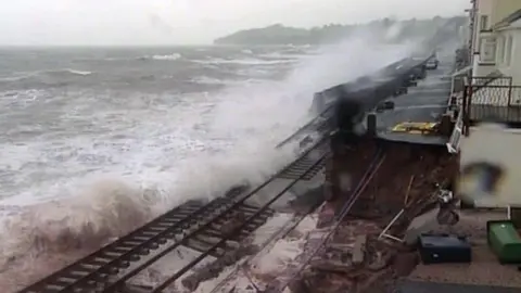 A damaged section of the coastal railway at Dawlish in 2014, with waves washing over broken track where the sea wall has collapsed. Houses sit close to the line on the right under a grey sky.