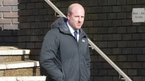 Eddie Mitchell A bald man in a black coat and navy suit walking down stairs and looking at the camera.