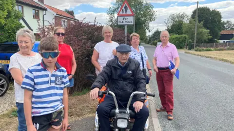 Owen Sennitt/BBC A group of people are standing next to the road. They include two men, one sitting on a mobility scooter, four women and a boy. There are homes and greenery in the background.