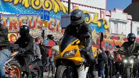 Several motorcyclists ride along Southend seafront past the arcades. All motorcyclists are wearing black helmets. The closest motorbike is mostly yellow in colour.