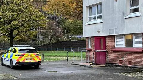 A police car parked outside Overtoun Court in Clydebank. The police car is white, with luminous yellow, red and blue detailing and 'Police' written on the side in blue text. The block of flats has exposed brick on the ground floor, with a red door and a window in the middle of two white grates. Above it, the building is light stone pebble dashed and has windows with white frames.