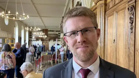 A man with blond hair and a short beard wearing glasses wears a dark suit and stands in a room filled with people over his right shoulder. 