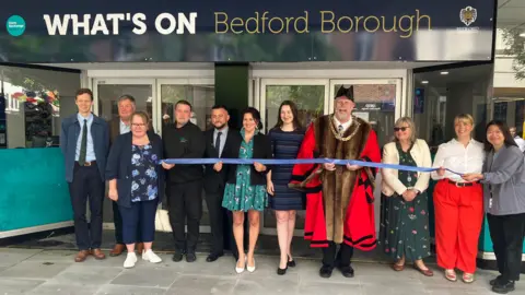 A picture of eleven people stood outside a building, including the Mayor of Bedford Tom Wootton, who is wearing his mayoral regalia. They are holding a ribbon.