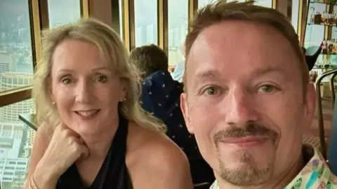 Warren Bullock Jane and Warren Bullock, a blonde woman in a black halter top is smiling at the camera next to a man with short light brown hair and a goatee