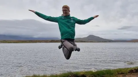 Tom Conroy Tom Conroy has been captured in the midst of a dancing move. He appears to be floating in mid air with his legs crossed and arms. Behind him is a vast lake and landscape settings. He has ginger hair, wearing glasses and is wearing a green jacket.