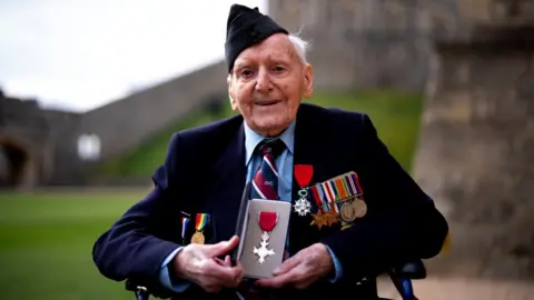 An elderly man wearing a military uniform with a hat and medals, holds his MBE medal in both hands and smiles while sitting in a wheelchair at Windsor Castle.