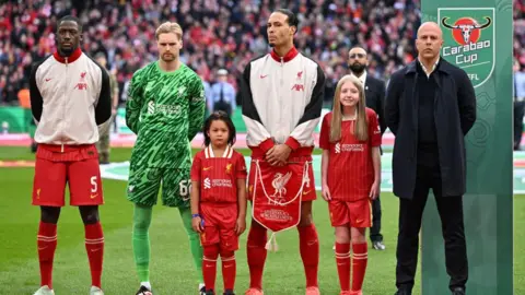 Getty Images Ibrahima Konate, Caoimhin Kelleher, Virgil van Dijk and Arne Slot, Manger of Liverpool, line up with their mascots prior to the Carabao Cup Final between Liverpool and Newcastle United at Wembley Stadium. Ibrahima Konate and Virgil van Dijk are wearing the Liverpool FC kits with bomber jackets, Caoimhin Kelleher is wearing a green goalkeeper kit. Arne Slot is wearing a black coat, black trouser and black half-zip with white shirt underneath. Hannah is in between Arne Slot and Virgil van Dijk.