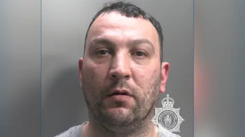 A mugshot of Edwards with a stubbly, black beard with short black hair. There is a North Wales Police logo in the bottom right-hand corner.