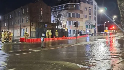 Water flows down a road. A handful of firefighters stand behind an orange barricade 
