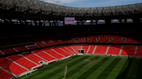 Reuters A view inside the Puskas Arena. Water sprinklers are spraying a green striped pitch. Around the ground are bright red seats, and most of the stadium is covered in shadow.