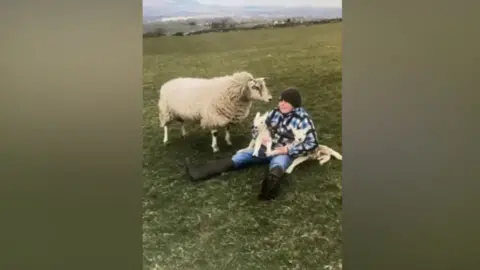 Hector Eccles sitting in a field next to a sheep. The boy wears a black beanie hat as well as a black, grey and white lumber jacket with blue jeans and black wellies.