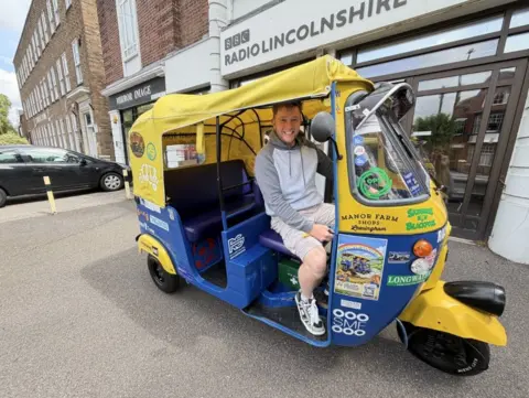 A man with a grey hoodie and shorts sits in a yellow and blue tuk-tuk.