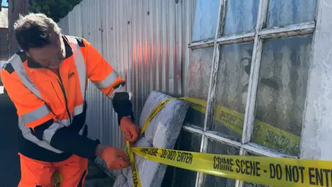 Gabby Colenso/BBC One of Luton's fly-tipping investigators, wearing orange protective clothing, ties yellow tape around dumped furniture.