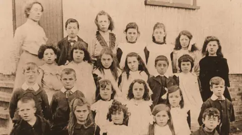 Walderslade Primary School A black and white photo showing 23 young pupils outside a school and an adult woman stood next to them