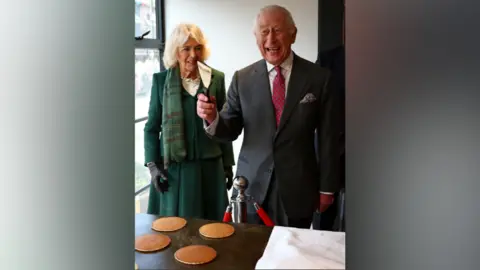 Reuters King Charles III uses a spatula during a tour of 200-year old "Hunters Bakery" during a visit with Britain's Queen Camilla to Limavady. Queen Camilla is beside him. The king has white hair and is wearing a dark grey suit, white shirt and red polka dot tie. The queen has blonde hair and is wearing a green skirt suit with green tartan scarf and black leather gloves. Four pancakes a on a table in front of them.